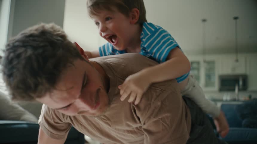 A man does pushups with a boy lying on his back in the living room of a house during the day