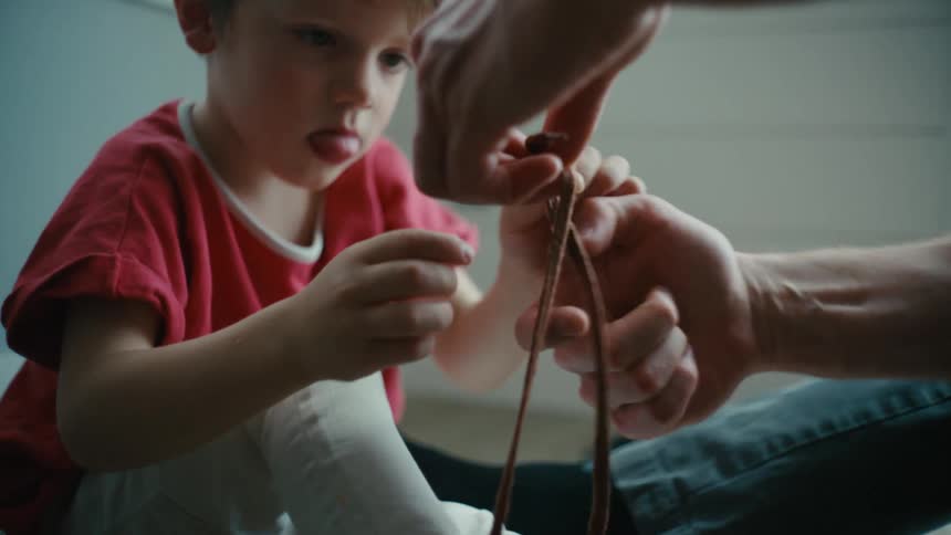 An adult helps a boy to tie his shoelaces while sitting on the floor indoors