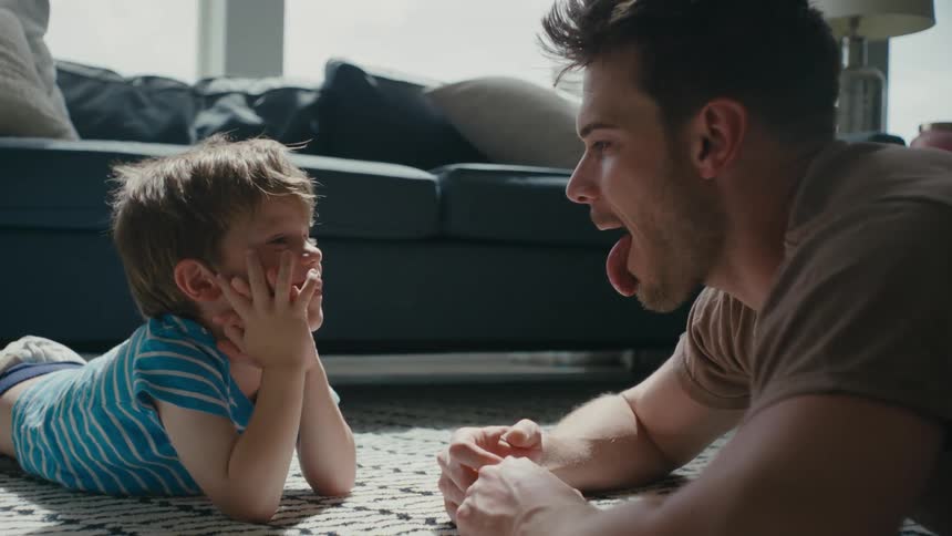 Father and son lying on the floor play with each other in the living room showing tongue