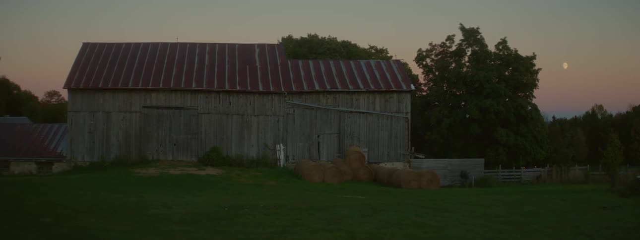 A barn structure with hay bales sitting outside trees behind the barn and the moon starting to come out at dusk