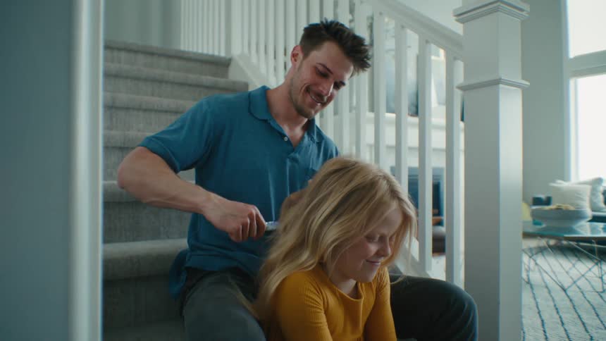 A man brushes the hair of a girl while they sit on the stairs in a house during the day