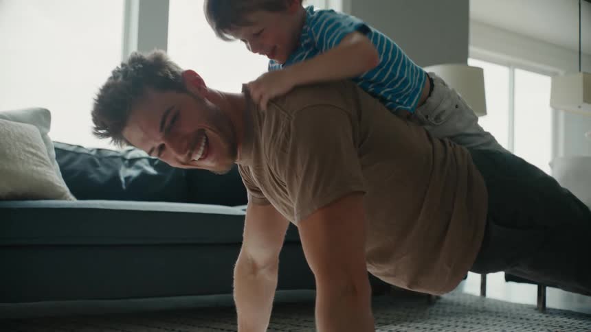 Father doing pushups on the living room floor plays with his son riding on his back