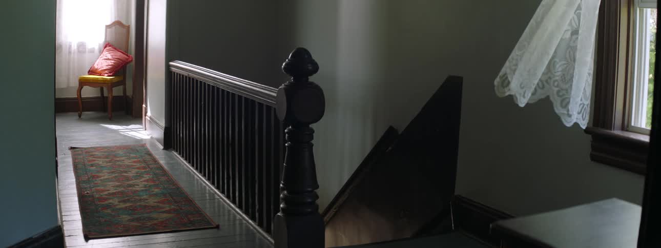 A hallway in the first floor of a house with wooden stairs going down and a curtain moving from the wind coming in from the window