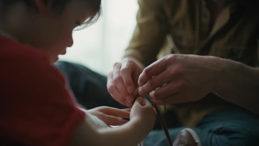 A man helps a boy to tie his shoes while they sit together on the floor during the day