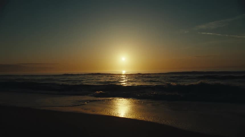 Sun shines from red horizon onto surf rolling onto beach