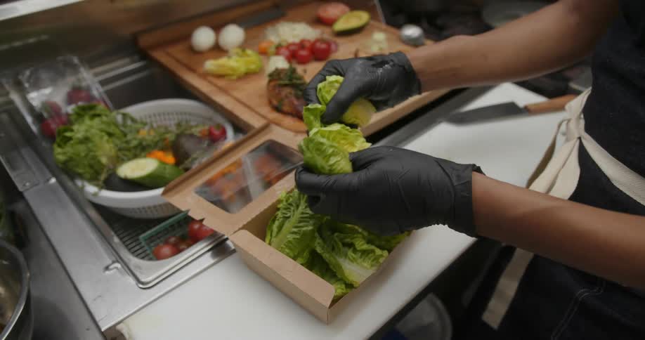 Female chef places lettuce in to go box while preparing salad near sink