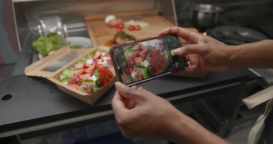 Chef standing in kitchen takes pictures of salad in a to go box with a smartphone