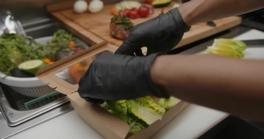 A pair of gloved hands prepares a lettuce based salad in a togo container