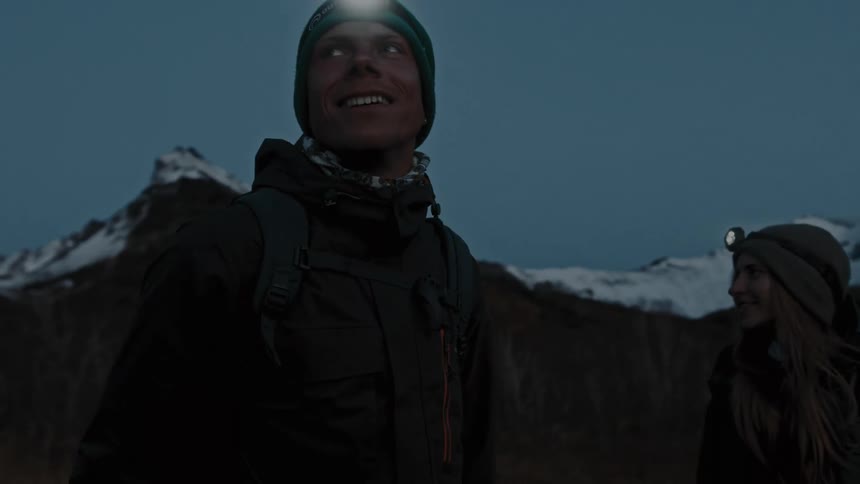 Two people are standing near mountains wearing winter coats and headlamps smiling as they look around