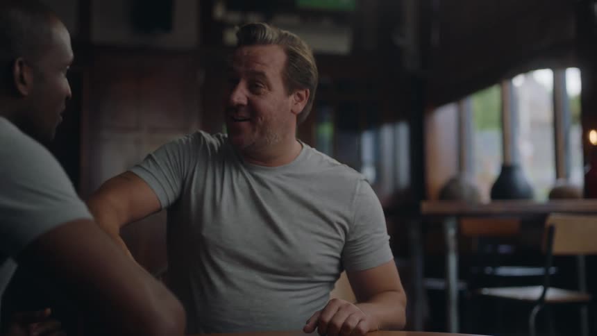 A man sitting at a table in a restaurant as two of his friends walk up behind him and they greet each other with smiles and handshakes