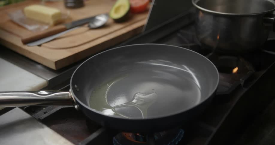 A person pours cooking oil into a frying pan on an oven in a kitchen