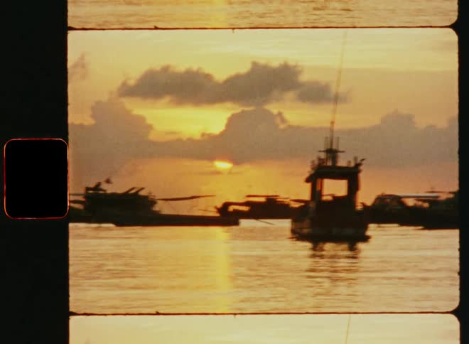 The silhouette of multiple ships docked in a harbor as the sun sets behind the clouds