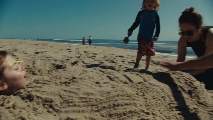 Boy buried in sand with his head sticking out while a little boy walks by with a shovel in his hand and stands near the mother