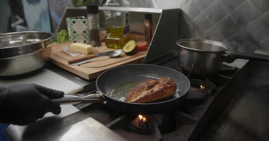 Person wearing black gloves cooking chicken in a frying pan on a stove