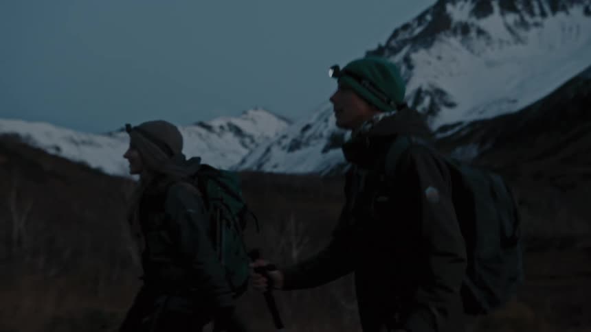 A pair of hikers cross a grassland beneath a snowy mountain range in the evening