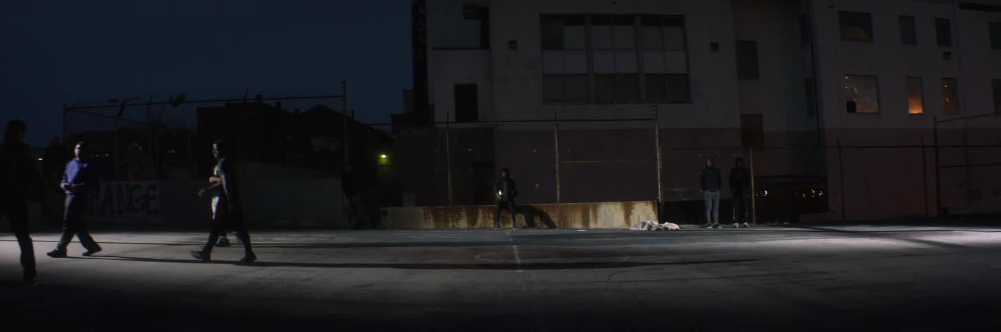 A young man is sitting on a fence watching others play basketball