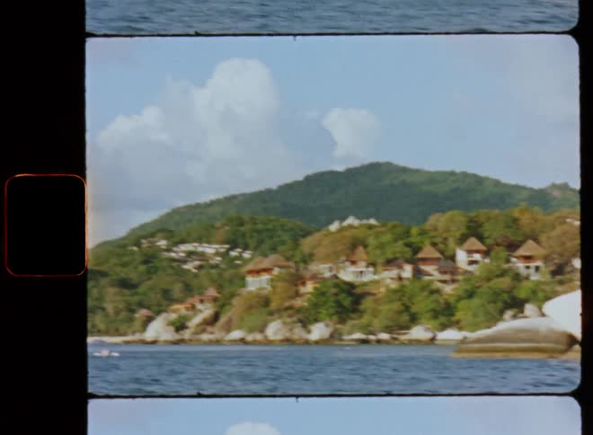Old film footage of houses on the coast and waves rippling in the water as a person paddles their boat