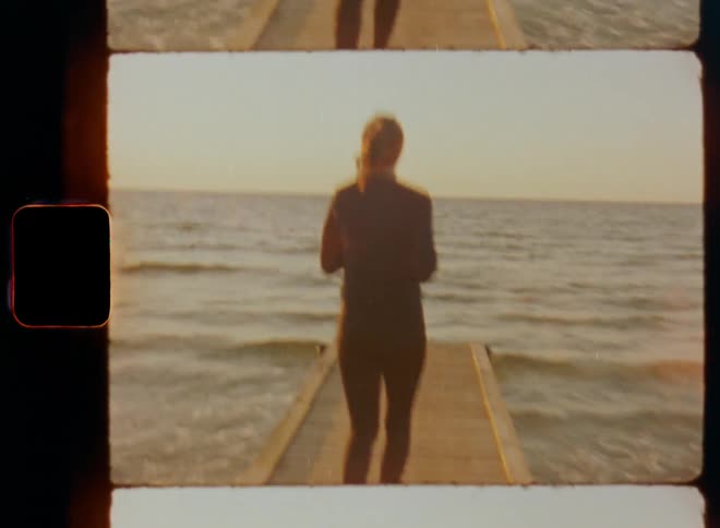 Attractive woman jogger runs or jogs along wooden pier at coast towards horizon as waves wash up onto the beach and the fresh coastal wind moves her brown hair in a ponytail during sunny day with blue sky and warm sunshine on vintage Super 8 mm film