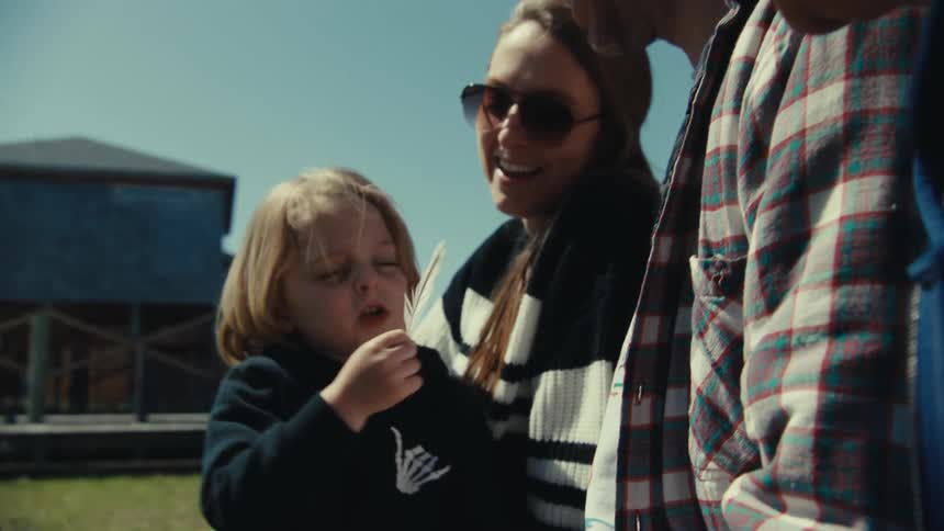 Kid shows feather to family as they sit outside across grass from building on stilts