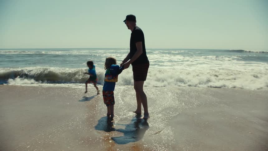 Father holding his sons hands as his other son runs around them in the water as the father lifts the other son as a wave comes ashore