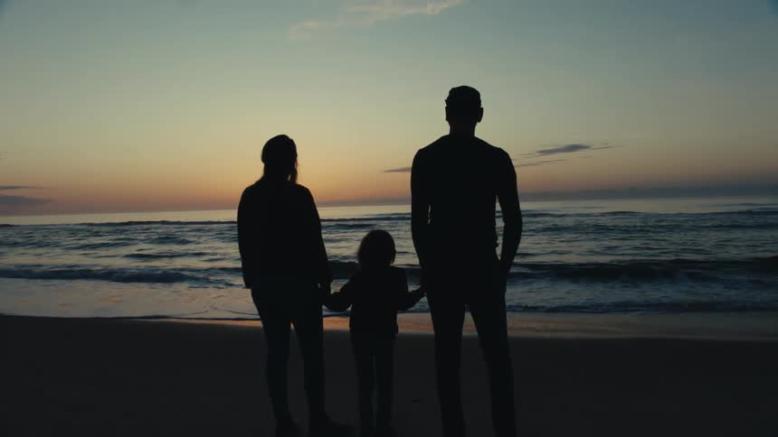 Silhouettes of a man woman and child holding hands on a beach looking out at the water