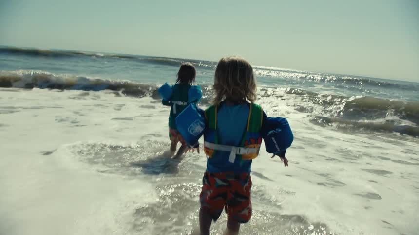 A pair of children wearing floatation devices play in the water on a beach during the day