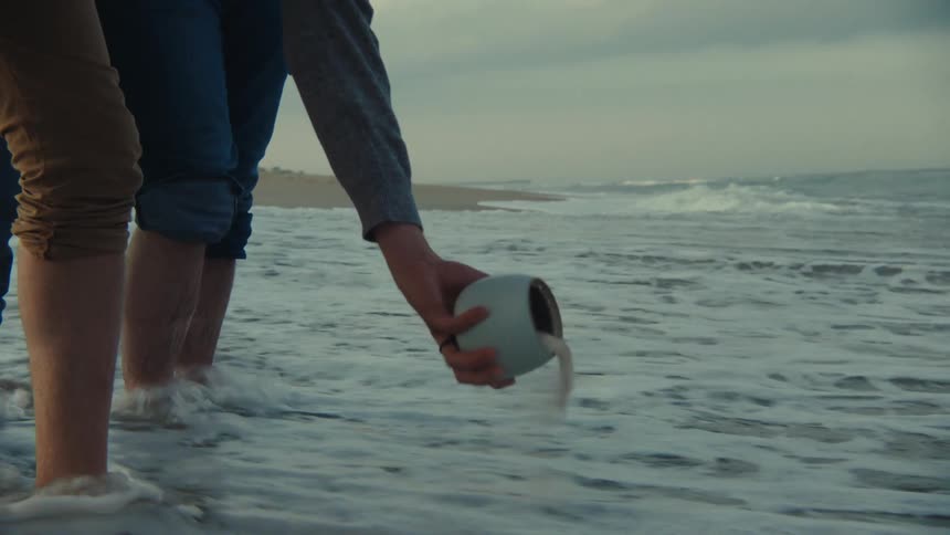 Two men in the beach pouring ash from a cup into the ocean