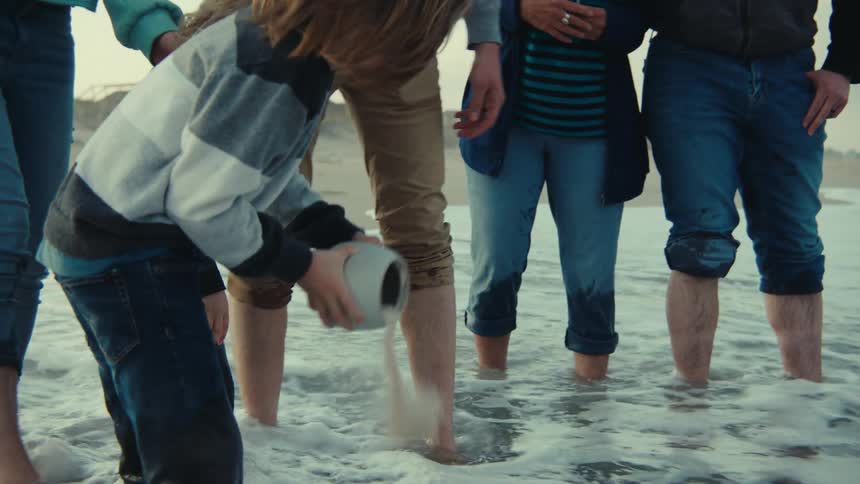 A family of men women and children standing on the beach as a child pours dust out of a jar into the water