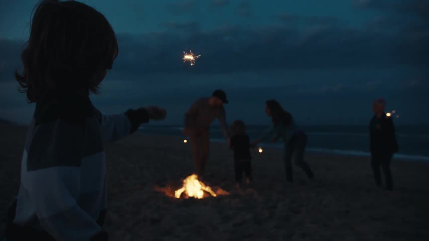 A family playing with sparklers on the beach next to a bonfire at night