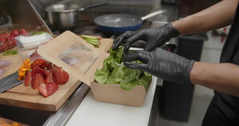 A close up of a persons hands wearing black gloves putting spinach into a food box next to fruit and other chopped up produce