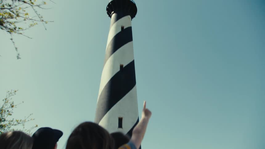 A mother and father carrying their children on their backs as they walk toward a lighthouse