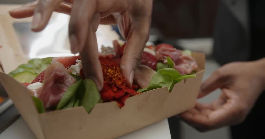 A close up of a salad prepared in a food box as a person places an edible flower in the box