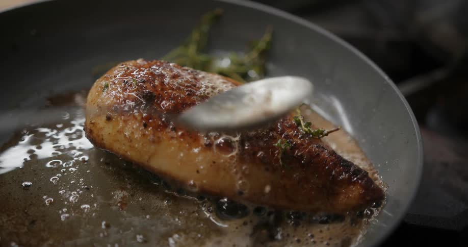 A fish fillet cooks on a frying pan as a spoon takes oil from the pan and spreads it over the fish