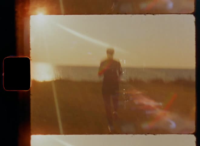 Handheld dynamic 8mm film footage with shaky camera movement of a female woman athlete wearing sporty fitness clothing and sneakers running on a green field with high grass close to the ocean into the sunrise on a sunny summer day