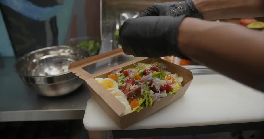 A person wearing black gloves placing food onto a prepared salad in a food box on the counter
