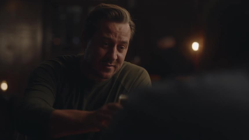 Man carrying beers towards a table with other people then sitting down and drinking with them