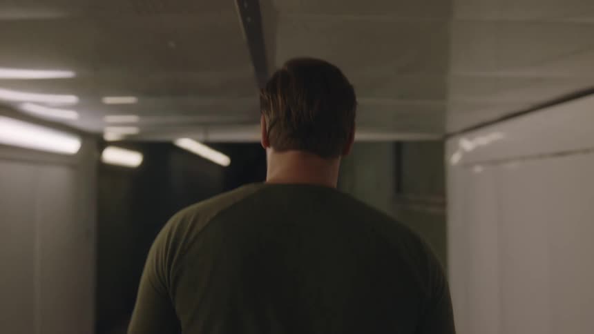 The backside of a man walking down a white lit corridor walking past another person walking as the man stops and starts to think