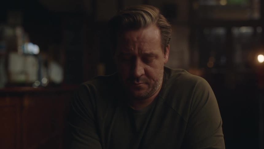 Three men sitting together drinking while one man is getting upset and red in the face looking down at the table