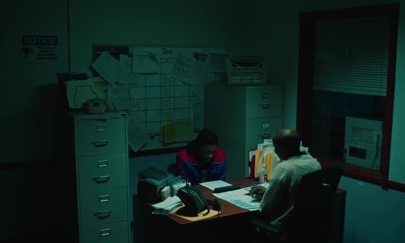 Teenage boy sitting across from a woman at her desk before they both stand up the boy revealing his handcuffs and they walk out of the room