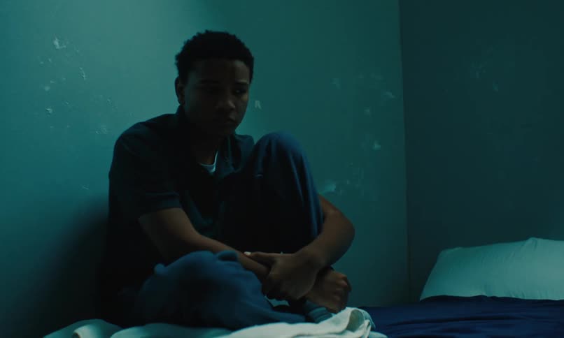 Youth frowns while sitting on bed against utilitarian walls with sticky tape residue
