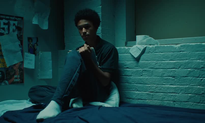 A teenage boy sitting on his bed writing in a notebook leaned against a brick wall