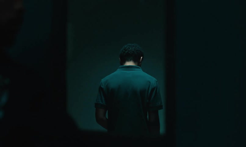 A policeman opens a door to a dark room leads a young man inside then comes out and locks the door the young man looking at the doors window with sadness