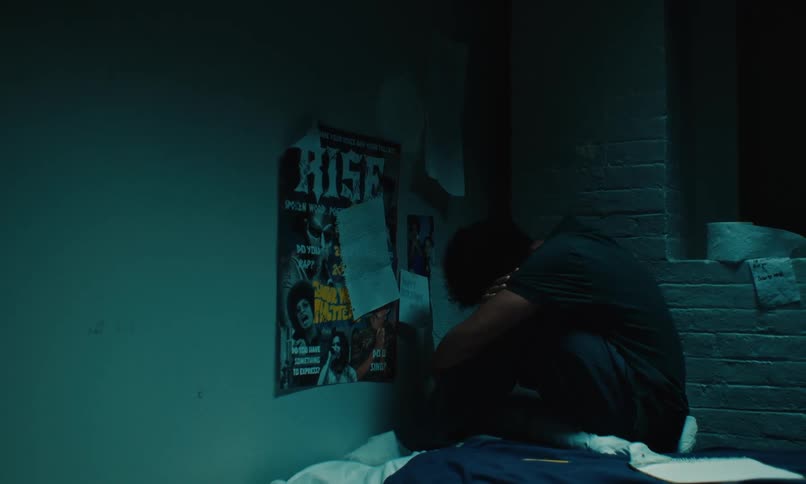 A young teenager sitting curled up on his bed next to a brick wall in front of posters on his wall