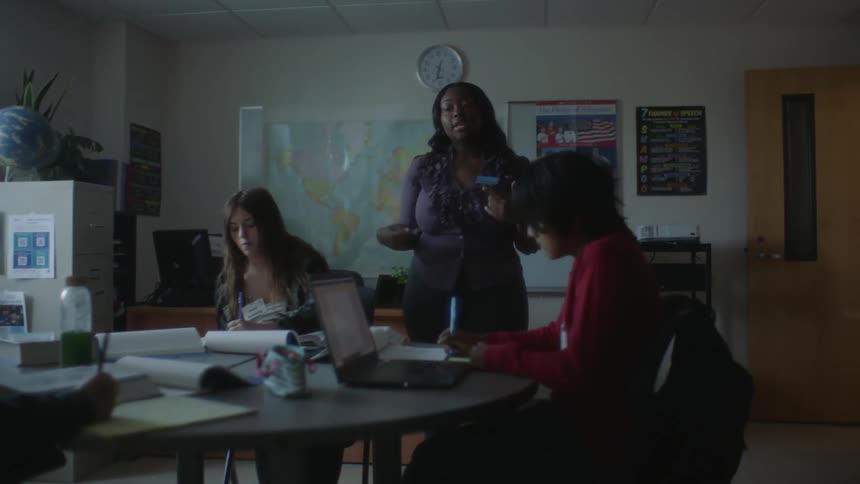 A teacher speaks to her students who sit at a desk and take notes