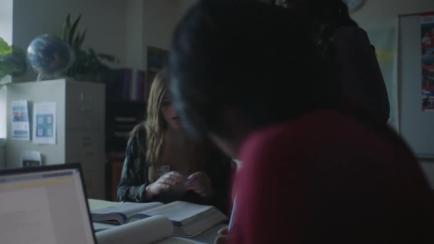 A teacher leaning over afemale student looking at a book while a male student writes on paper next to them