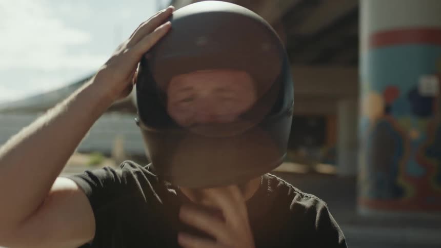 Close up of a man putting on a helmet while sitting in front of a bridge with tall pillars