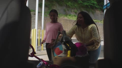 A pair of women retrieve a box and a bicycle for children from the back of a vehicle during the day