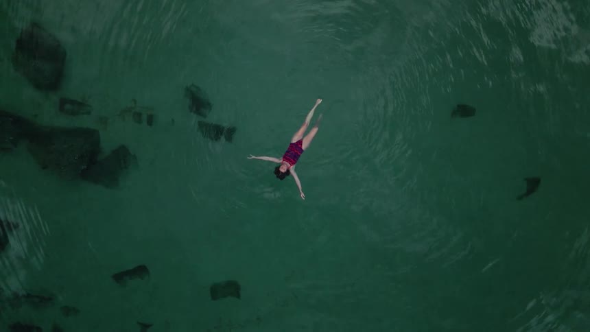 An aerial straight down of a woman swimming on her back in a clear body of water