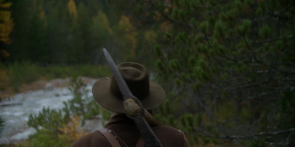 Miner walks along trail through woods toward river while carrying pickax and a mining pan