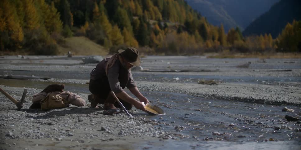 An older man sifting rocks in a pan next to a river with a forest and mountains in the background